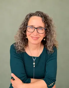 Lucrezia Mangione smiling, wearing glasses and a deep teal top against a soft sage background.
